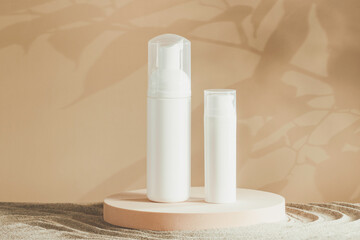 Two white blank plastic packaging cosmetic pump bottles for mousse foam cream and lotion stand on round podium, beach sand and beige background. Front view