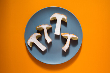 Wall Mural - Halfs, sliced portions of fresh porcini mushrooms in grey round plate on orange surface close-up, top view