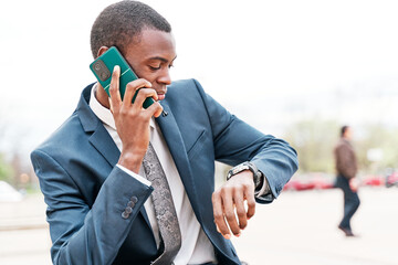 Wall Mural - Handsome man having a conversation on smart phone arranging a meeting looking at wrist watch to be on time. Young African American businessman in suit and tie.