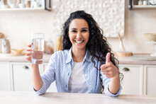 Excited Latin Woman Holding Glass With Clean Water And Showing Thumb Up, Sitting At Table In Kitchen Interior
