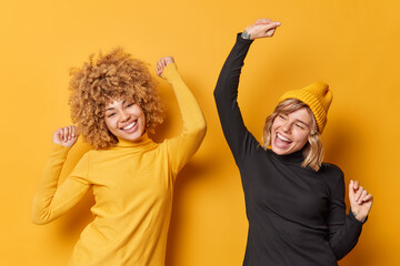 Wall Mural - Positive cheerful young women have fun and dance carefree shake arms dressed in casual turtlenecks have glad expressions isolated over yellow background. People happiness and emotions concept