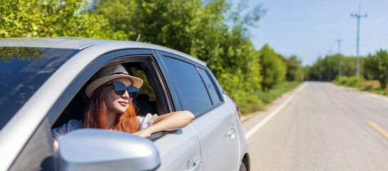 Woman with ginger red hair wearing sunglasses driving car in solo weekend getaway vacation trip looking out the window in summer with lush green nature in countryside road for freedom and adventure