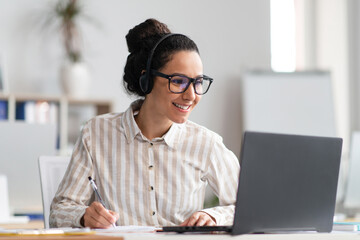 Poster - Portrait of happy young latin woman wearing wireless headphones watching online course, sitting at table