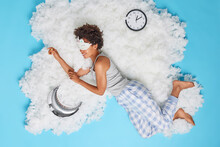 Positive Relaxed Woman Takes Nap During Daytime Poses Bare Feet In Pajama Smiles Happily Wears Sleepmask On Eyes Poses On White Cloud With Inflated Moon And Clock Showing Time. Blue Background
