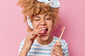 Wall Mural - Positive woman with curly hair eats delicious macaroon doesnt care about teeth health holds wooden toothbrush winks eye enjoys sweet tasty food isolated over pink background. Hygiene of oral cavity