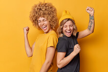Two Positive Women Friends Feel Very Happy Like Winners Make Triumph Gesture Shake Arms Celebrate Success Stand Back To Each Other Dressed In Casual T Shirts Isolated Over Yellow Background.