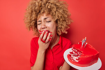 Wall Mural - Crying dissatisfied curly haired young woman eats festive cake with doleful expression feels lonely and unnecessary on Valentines Day poses against vivid red background. Bad mood during holiday