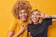 Positive women have joyful expressions smile broadly make peace gesture laugh at something funny foolish around dressed casually isolated over vivid yellow background. Happy emotions concept
