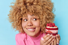 Curly Haired Beautiful Woman Bites Lips Looks At Appetizing Piece Of Cake With Raspberries Has Sweet Tooth Feels Temptation To Eat Delicious Dessert Isolated Over Blue Background. Sugar Addiction