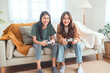 Two happy asian roommates sitting on couch in living room at home enjoy and excited holding console playing game together.