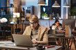 © Mediaphotos - Young Caucasian man in eyeglasses and jacket sitting at desk and using laptop while working In Dark Office