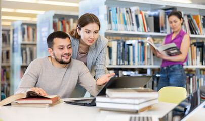 Wall Mural - Young positive interested couple of students spending time together in library, reading books and browsing educational websites on laptop