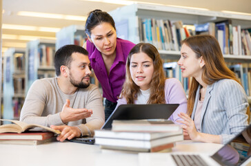 Wall Mural - Cheerful young adult university students studying in library using books and laptop, talking and smiling while working on group project. Brainstorming and teamwork concept