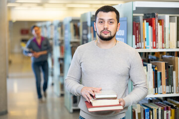 Portrait of confident successful young adult bearded student holding stack of books, standing in university library and looking at camera..