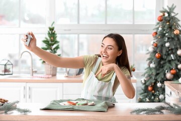 Wall Mural - Beautiful housewife near plate with gingerbread cookies taking selfie in kitchen