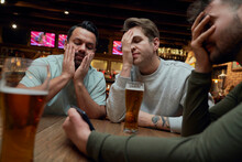 Three Frustrated Male Soccer Fans Having Beer And Watching A Match On Smartphone In A Pub