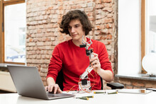 Smiling Teenage Boy With Robotic Arm Using Laptop Sitting At Table