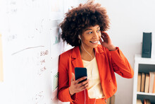 Smiling Businesswoman Holding Smart Phone Listening Music Through In-ear Headphones At Office
