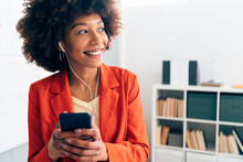 Happy Businesswoman Holding Mobile Phone Listening Music Through In-ear Headphones In Office