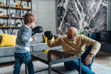 Playful Grandfather And Grandson Wearing Boxing Glove Playing In Living Room At Home