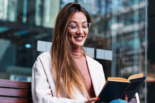 Smiling Woman Wearing Eyeglasses Reading Book Sitting In Front Of Glass Wall