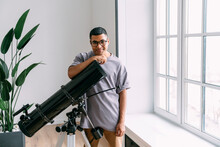 Smiling Man Wearing Eyeglasses Leaning On Telescope Standing By Window At Home