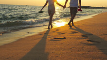 Couple Walking Together On The Beach At Sunset, Young Happy Couple Holding Hands Walking Along Beach, Walking Barefoot And Carrying Shoes, Outdoor Leisure Time By The Seaside.