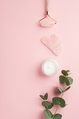 Wall Mural - Skincare concept. Top view vertical photo of rose quartz roller gua sha cream bottle and eucalyptus sprig on isolated pastel pink background