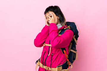 Wall Mural - Young mountaineer girl with a big backpack over isolated pink background covering eyes and looking through fingers