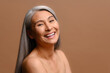 © Vadim Pastuh - Asian mature magnificent grey-haired woman isolated on brown background looking at camera and smiling toothy. Well-looking middle-aged lady laughing playfully, hand near face. Anti-aging care