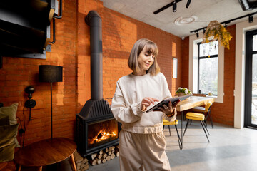 Portrait of a young pretty woman standing with a digital tablet in cozy living room. Interior in loft style with burning fireplace on background. Idea of smart home or work from home