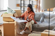 © Syda Productions - moving, people and real estate concept - happy smiling woman with blueprint and boxes sitting on sofa at new home