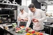 © AnnaStills - Young chef in uniform choosing fresh vegetables for dish at table with his assistant making notes on paper, they working in commercial kitchen