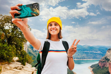 Wall Mural - Portrait of young cheerful woman backpacker with short brunette hair and yellow beanie, taking selfie with smartphone doing victory sign with hand over the top of the mountain lake. Adventure, travel.