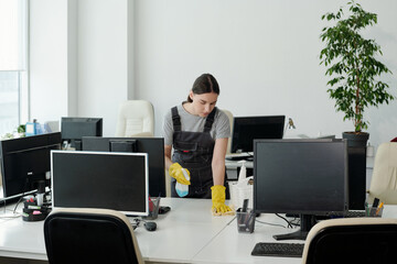 Poster - Young woman in coveralls and gloves using detergent while wiping one of desks with computer monitors in large openspace office