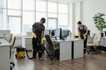 Poster - Young black man in workwear cleaning floor while girl wiping desks with computer monitors in large contemporary openspace office