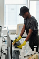 Poster - African American male staff of cleaning company in black t-shirt, cap and coveralls and yellow gloves wiping computer keyboard in office