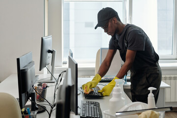Poster - Young contemporary black man in uniform cleaning keyboard of computer with duster and detergent in openspace office