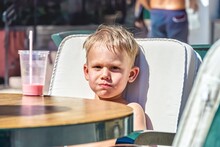 Cute Blond Boy Sits In Chair At Table Drinking Milkshake In Street Cafe Near Beach In Omis. Toddler Enjoys Vacation In Croatia On Sunny Day Closeup