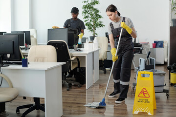 Poster - Young black man wiping computer monitors while Caucasian woman in coveralls and yellow gloves with mop cleaning floor in office