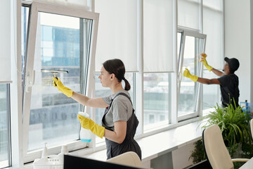 Poster - Young female cleaner in workwear washing large windows of openspace office against her African American male colleague