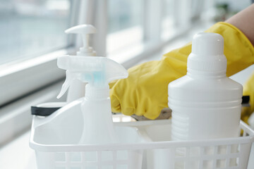Poster - Group of white plastic bottles with detergents for washing windows in container against hands of female cleaner in yellow gloves