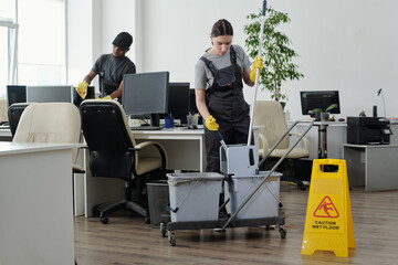 Poster - Young cleaning lady sqeezing mop in bucket against male colleague while going to wash floor in large modern openspace office