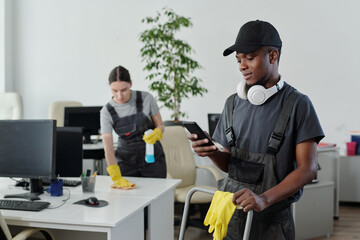 Poster - Young male cleaner in workwear choosing music in playlist in his smartphone during work in contemporary openspace office