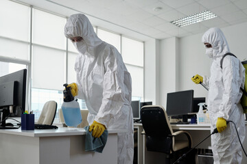Poster - Workers of cleaning service in hazmat suits disinfecting furniture and computer equipment in large modern openspace office