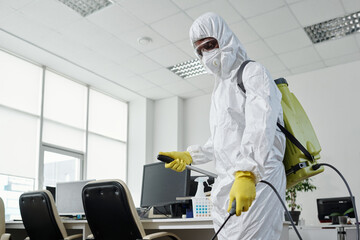 Poster - Young black man in hazmat suit spraying floor, furniture and other equipment in modern openspace office with disinfectant