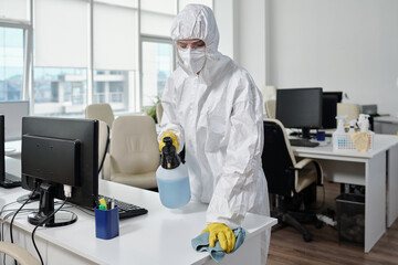 Poster - Young female worker of cleaning service in hazmat suit wiping surfaces of desks and computer equipment in modern openspace office