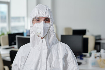 Poster - Young woman in white overalls, respirator and protective eyeglasses looking at camera while working in modern openspace office