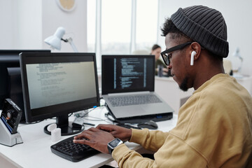 Poster - Side view of young male programmer or it developer in casualwear listening to music in earphones while working in office