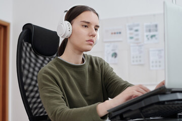 Poster - Young serious female programmer in headphones and casualwear decoding data in front of computer monitor by her workplace